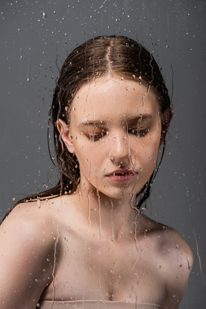 Fair haired model with naked shoulders standing behind wet glass on grey background  - Photo, Image