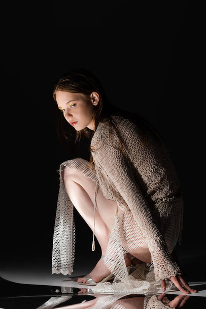 Stylish young woman in fishnet skirt and knitted cardigan posing near reflective surface on black background  - 写真・画像