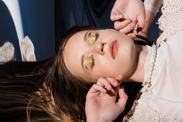 Horní pohled na ženu se zlatým make-upem ležící na reflexním povrchu na abstraktním pozadí  - Fotografie, Obrázek