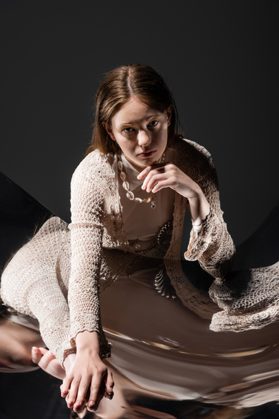 Stylish young woman in seashell necklace and knitted cardigan touching reflective surface on grey background  - 写真・画像