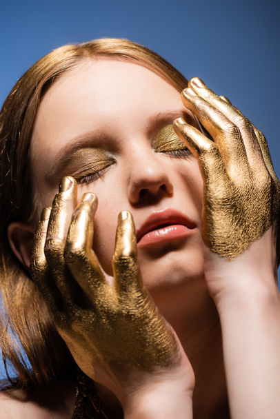 Portrait of young woman with makeup and golden paint on hands touching face isolated on blue  - Fotografie, Obrázek