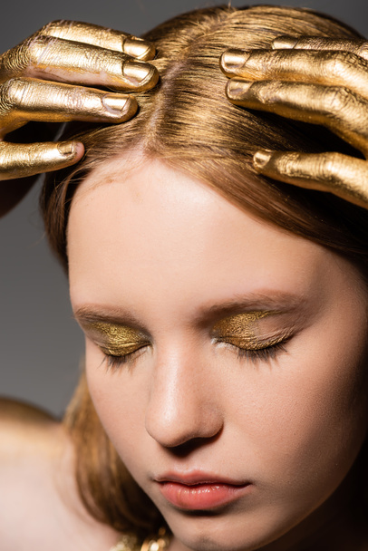 Portrait of young woman with golden makeup and paint on hands touching hair and closing eyes isolated on grey  - Fotoğraf, Görsel