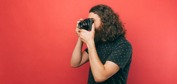 Boční pohled fotografie vousatý hispter muž s dlouhými vlasy dělat fotografii s kamerou přes růžové pozadí. - Fotografie, Obrázek