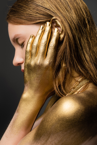 Side view of young woman in golden paint touching ear isolated on grey  - Photo, Image