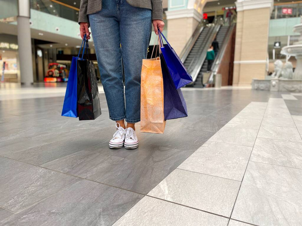 手に持っている認識できない女性の着用ジーンズパンツ購入の完全な多くのショッピングバッグ。紙袋でショッピングモールの真ん中に立っている女性の足の閉じる。販売コンセプト - 写真・画像