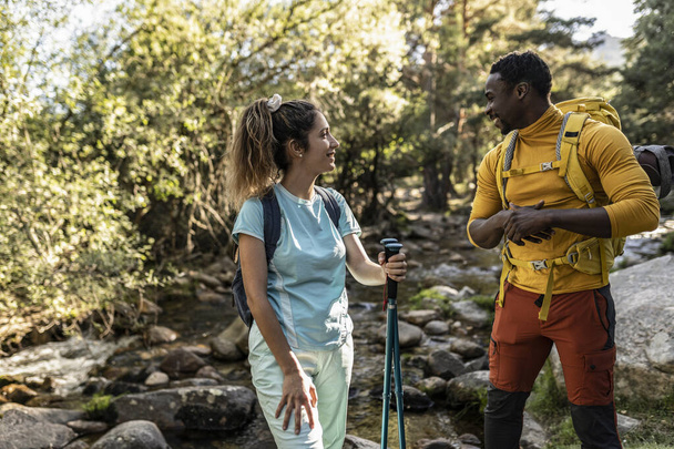 Multiracial hiker couple with backpackers hiking in the wood with the help of hiking poles. Nature and environmental themes - Photo, Image