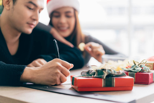 Κοντινό πλάνο το χέρι του ανθρώπου γράφει σε μια κενή χριστουγεννιάτικη κάρτα με ένα στυλό. Ζευγάρι κάθεται και γράφει χριστουγεννιάτικη κάρτα μαζί για την αποστολή με δώρα έκπληξη στο σπίτι κατά τη διάρκεια των διακοπών των Χριστουγέννων. - Φωτογραφία, εικόνα