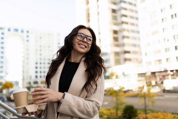 стильная и довольная деловая женщина с чашками кофе в руках на фоне городского пейзажа. - Фото, изображение