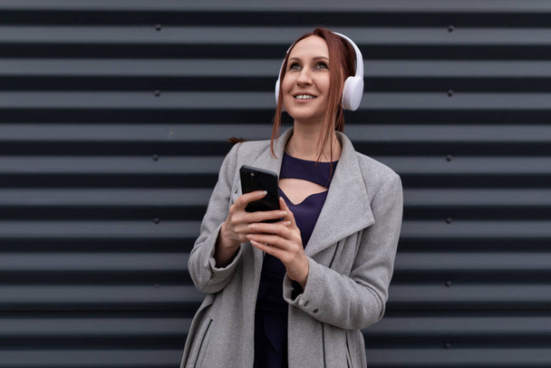 erwachsene rothaarige Geschäftsfrau auf dem Hintergrund einer grauen Wand mit Kopfhörern hört Musik am Telefon. - Foto, Bild