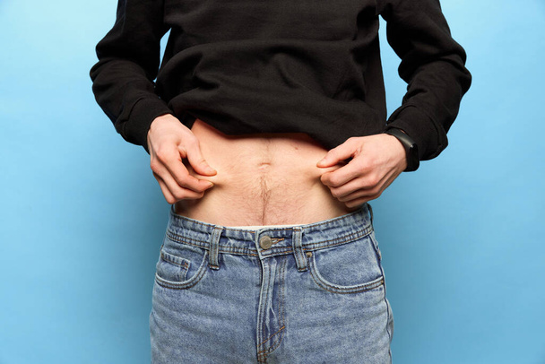Closeup male hands stretching skin on his belly, man showing to camera his abdomen. Concept of dieting, sport, fitness, healthy lifestyle, body positive and wellbeing. Liposuction and plastic surgery - Zdjęcie, obraz