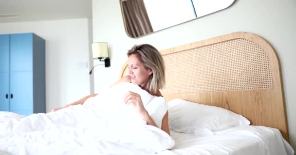 Šťastná ospalá krásná dospělá žena se probudí ležící v pohodlné bílé posteli. Pozitivní dívka protahuje těší brzy dobré ráno v útulné ložnici - Záběry, video