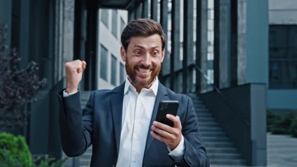 Ενθουσιασμένος έκπληκτος άνθρωπος στέκεται σε εξωτερικούς χώρους κοιτάζει την οθόνη του τηλεφώνου ανάγνωση καλά νέα γιορτάζει τη νίκη χαρούμενος ενθουσιώδης επιχειρηματίας νικητής χαίρεται για την επιτυχία λαμβάνει επιτυχημένο μήνυμα στο smartphone - Πλάνα, βίντεο
