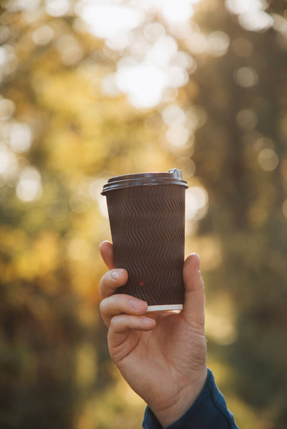 Una tazza di carta nera con caffè in una mano mans primo piano sullo sfondo del sole al tramonto tra le foglie degli alberi. Vista frontale - Foto, immagini