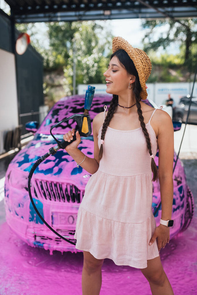 Young woman high pressure hose stands in front of a car covered in pink foam - Fotografie, Obrázek