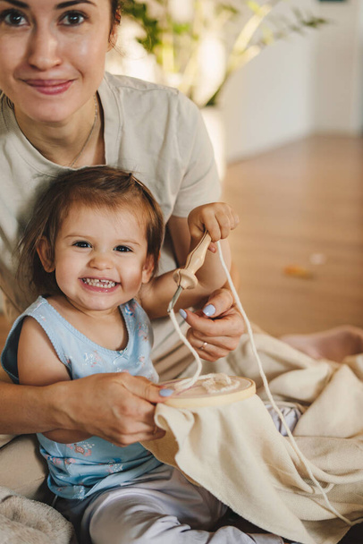 Primer plano de una madre con su bebé cosiendo artesanía bordado hecho a mano con accesorios e instrucción en casa sentado en el suelo. Educación familiar - Foto, imagen