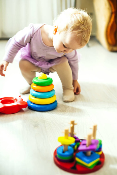 girl plays logical pyramid on floor in living room on sunny day. Montessori wooden toy folded pyramid. Circle, quadra, triangle, rectangle wooden elements of children's toys. Multi-colored toy blue, yellow, red, green - Foto, imagen