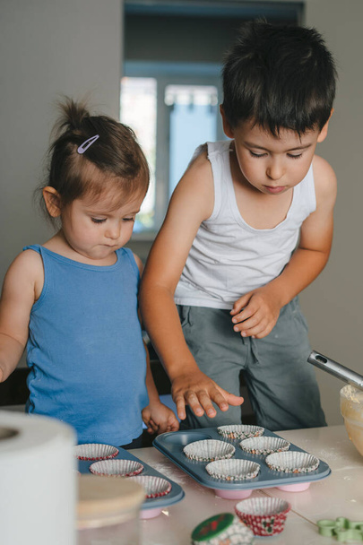 Two little caucasian kids filling the dough into cupcake silicone molds in the kitchen. Concept of confectionery cooking. Muffins making. Raw dough with berries - Fotoğraf, Görsel