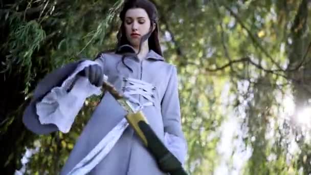 Mujer joven en la imagen de guerrero antiguo se encuentra bajo el árbol, toma su espada de vaina por las manos en guantes y luego va en el bosque. - Metraje, vídeo