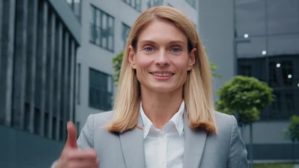 Close-up young smiling businesswoman in suit standing outdoors against backdrop office building looking at camera showing thumb up gesture approval sign achievement agreement symbol shows good result - Footage, Video