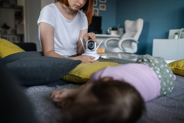 home surveillance camera unknown woman caucasian mother adjusting and setting up security equipment in order to monitor her baby child sleeping on the bed motherhood parenting concept focus on camera - Valokuva, kuva