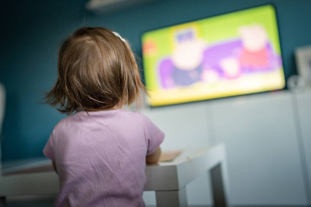 Вид ззаду на одну невідому кавказьку дівчинку-малятку, яка стоїть вдома, дивлячись телевізор, коли їсть копію простору дитинства, розвиваючи концепцію дозвілля
 - Фото, зображення