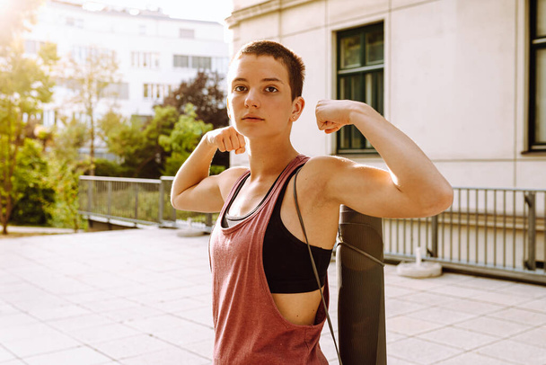 teenager girl with short haircut in sportswear demonstrates muscles of pumped up arms, sunny, morning or evening. Healthy lifestyle, sports, outdoor fitness. Useful training, athletic physique, strong muscles. - Photo, Image
