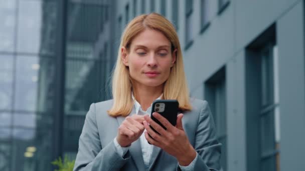 Caucasian businesswoman holding phone stands outdoors enthusiastic happy woman pointing at smartphone screen getting good news receives profitable business offer winning in online betting rejoices win - Footage, Video