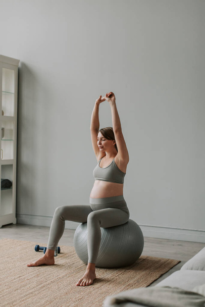 Mulher grávida atraente fazendo exercícios sentados na bola de fitness durante o treinamento na sala de estar em casa. Jovem grávida mãe tem treino de ioga. Conceito de gravidez  - Foto, Imagem