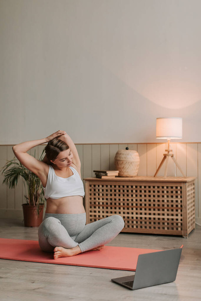 Junge Frauen dehnen sich zu Hause. Junge Frau macht Yoga auf Gymnastikmatte während ihrer Schwangerschaft im Wohnzimmer. Gesunde zukünftige Single-Mütter trainieren in Online-Kursen. Stretching, Schwangerschaftsyoga-Konzept - Foto, Bild