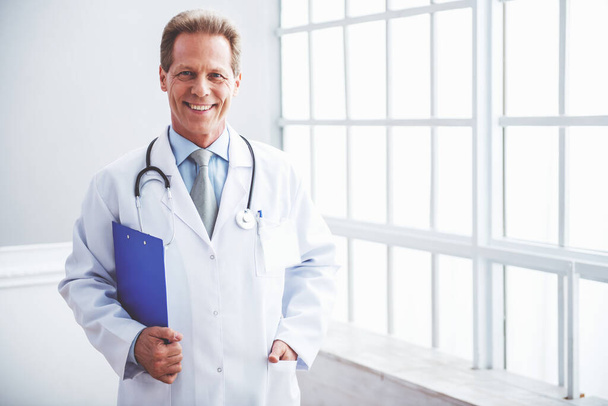 Komea keski-ikäinen lääkäri valkoisessa lääketieteellisessä takissa katsoo kameraa ja hymyilee, seisoo kansio käsissään.
 - Valokuva, kuva