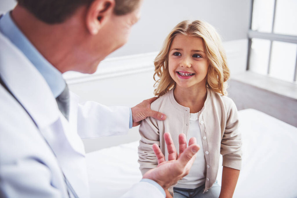 Χαριτωμένο μικρό κορίτσι ακούει παιδίατρος και το χαμόγελο, κάθεται στο γραφείο ιατρών - Φωτογραφία, εικόνα
