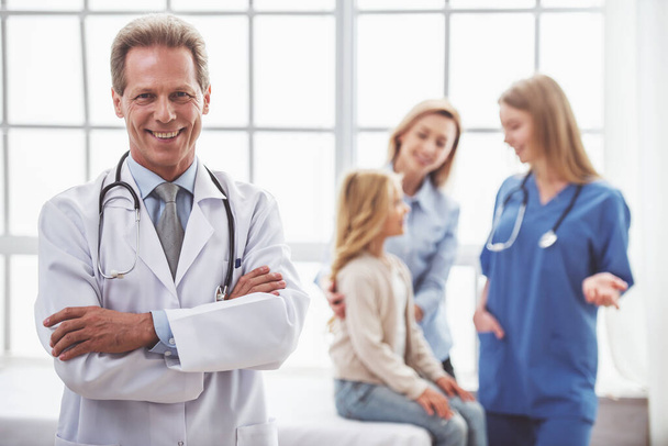 Όμορφος μέσης ηλικίας παιδίατρος σε λευκό παλτό στέκεται με σταυρωμένα χέρια και να χαμογελά, στο παρασκήνιο νεαρός γιατρός μιλάει στους ασθενείς - Φωτογραφία, εικόνα