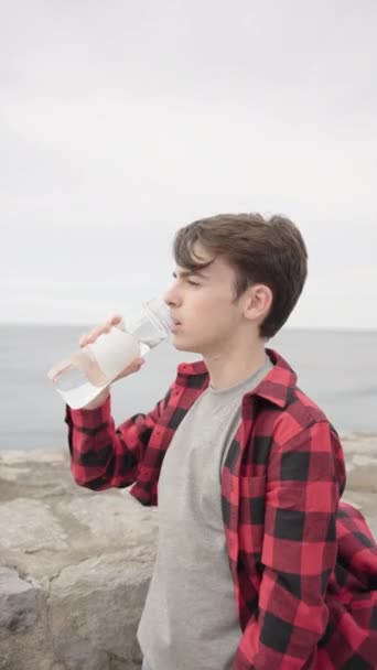 Jóképű tinédzser fiú vizet iszik egy üvegből a szabadban. Egészséges életmód és hidratációs koncepció - Felvétel, videó