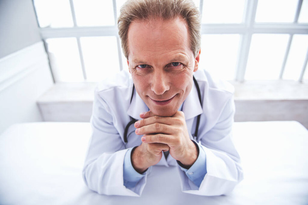 Vista de ángulo alto del guapo médico de mediana edad en bata médica blanca mirando a la cámara y sonriendo, apoyado en sus manos
 - Foto, Imagen