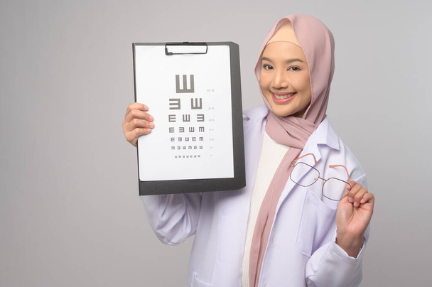 A Young muslim female ophthalmologist with glasses holding eye chart over blue background studio, healthcare concept - Photo, image