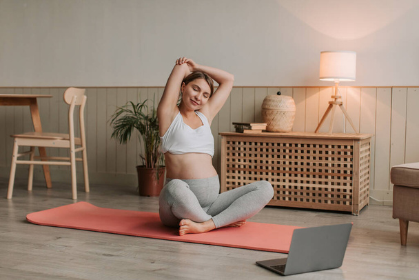 İnternette yoga yapan gülümseyen hamile bir kadın bilgisayara bakıyor. Hamilelik boyunca sağlıklı yaşam tarzı, sağlık ve spor kavramı. Yüksek kalite 4k görüntü - Fotoğraf, Görsel