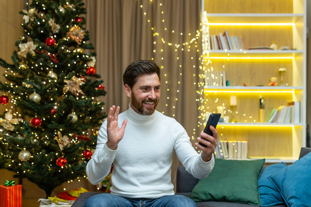 Businessman at home for Christmas and New Year holidays sitting on sofa in living room near Christmas tree talking on video call, man having fun talking with friends holding phone - Photo, image
