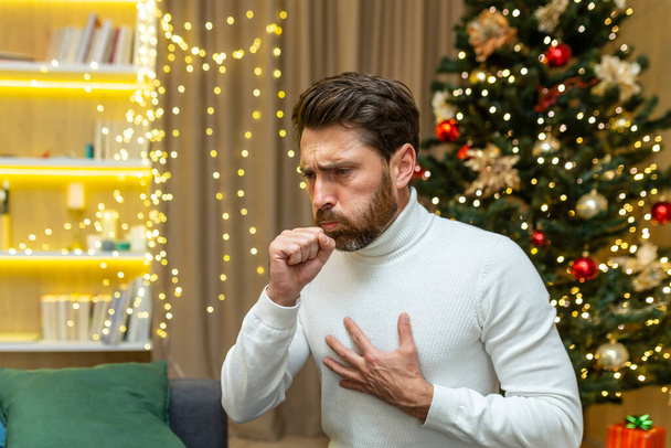 man coughs on christmas at home sitting alone on sofa sick on new year holidays in living room. - Φωτογραφία, εικόνα