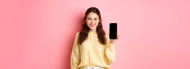 Technology and online shopping. Young smiling woman looking determined, advicing download app, showing smartphone app on screen, standing against pink background. - Foto, Bild