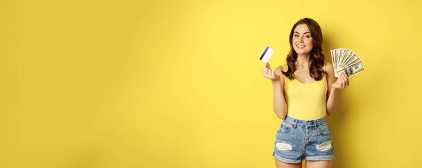 Young pretty woman in summer outfit holding credit card and money, cash in hands, standing against yellow background. - Foto, Imagen