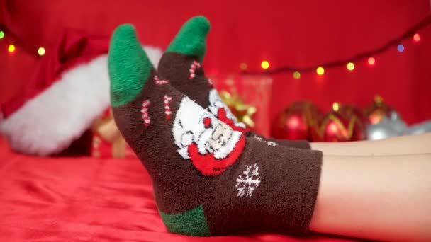 Pies en cálidos calcetines de Navidad con Santa Claus sobre un fondo rojo, esperando las vacaciones. - Imágenes, Vídeo
