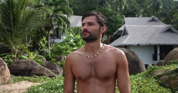 Atractivo hombre barbudo con torso desnudo contra casas en la selva tropical, disfruta tomando el sol, sonriendo hermosa sonrisa dentada - Imágenes, Vídeo