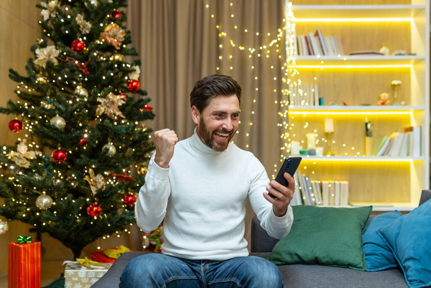 Man celebrating victory and success received good notification online message sitting on sofa near tree during christmas and new year holidays holding hand up success gesture using phone. - Zdjęcie, obraz
