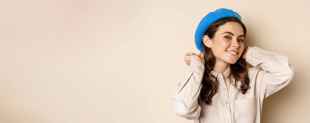 Stylish modern girl put on trendy hat on head and smiling, going out, posing against beige background. - Photo, Image