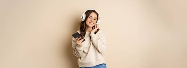 Happy caucasian woman listening music in headphones on smartphone app, dancing and having fun, standing over beige background. - Photo, image