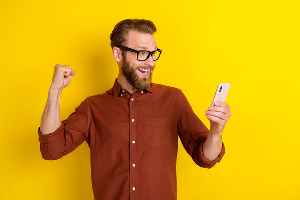 Photo of handsome man with blond beard dressed burgundy shirt clench fist impressed look at phone isolated on yellow color background. - Фото, зображення