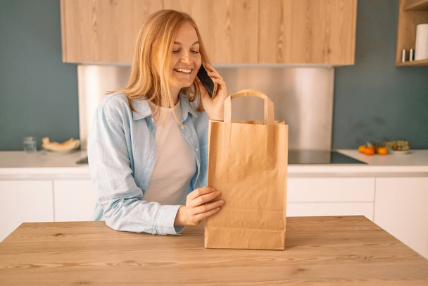 Mooie jonge blanke vrouw houden eco boodschappentas na het winkelen met behulp van smartphone in de moderne keuken, bezorgservice, huis aankoop in covid-19 lockdown.  - Foto, afbeelding