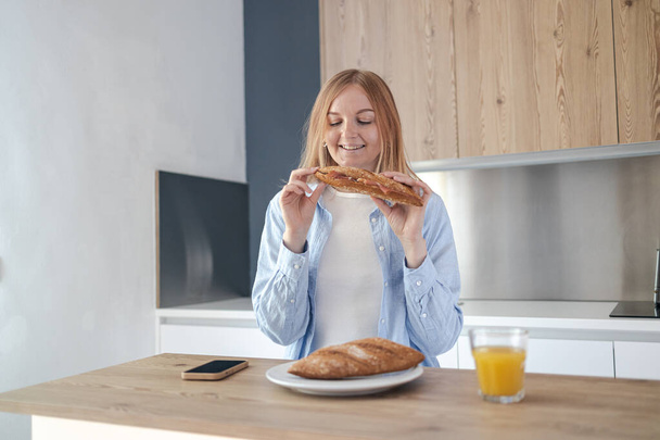 Close-up portrait of woman is in a hurry eats freshly baked baguette with prosciutto for breakfast in morning in light white interior style kitchen.  - 写真・画像