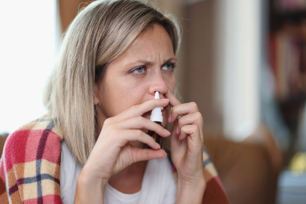 Porträt einer kranken Frau, die mit Nasentropfen in eine Decke gehüllt auf einem Sofa sitzt. Saisonale Krankheiten, Behandlungs- und Gesundheitskonzept - Foto, Bild