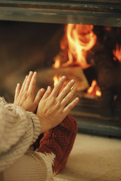 Femme en chaussettes de laine confortables réchauffer les pieds et les mains à la cheminée dans la pièce rustique. Maison chauffante en hiver avec poêle à bois. Jeune femme élégante assise à la cheminée dans une ferme - Photo, image